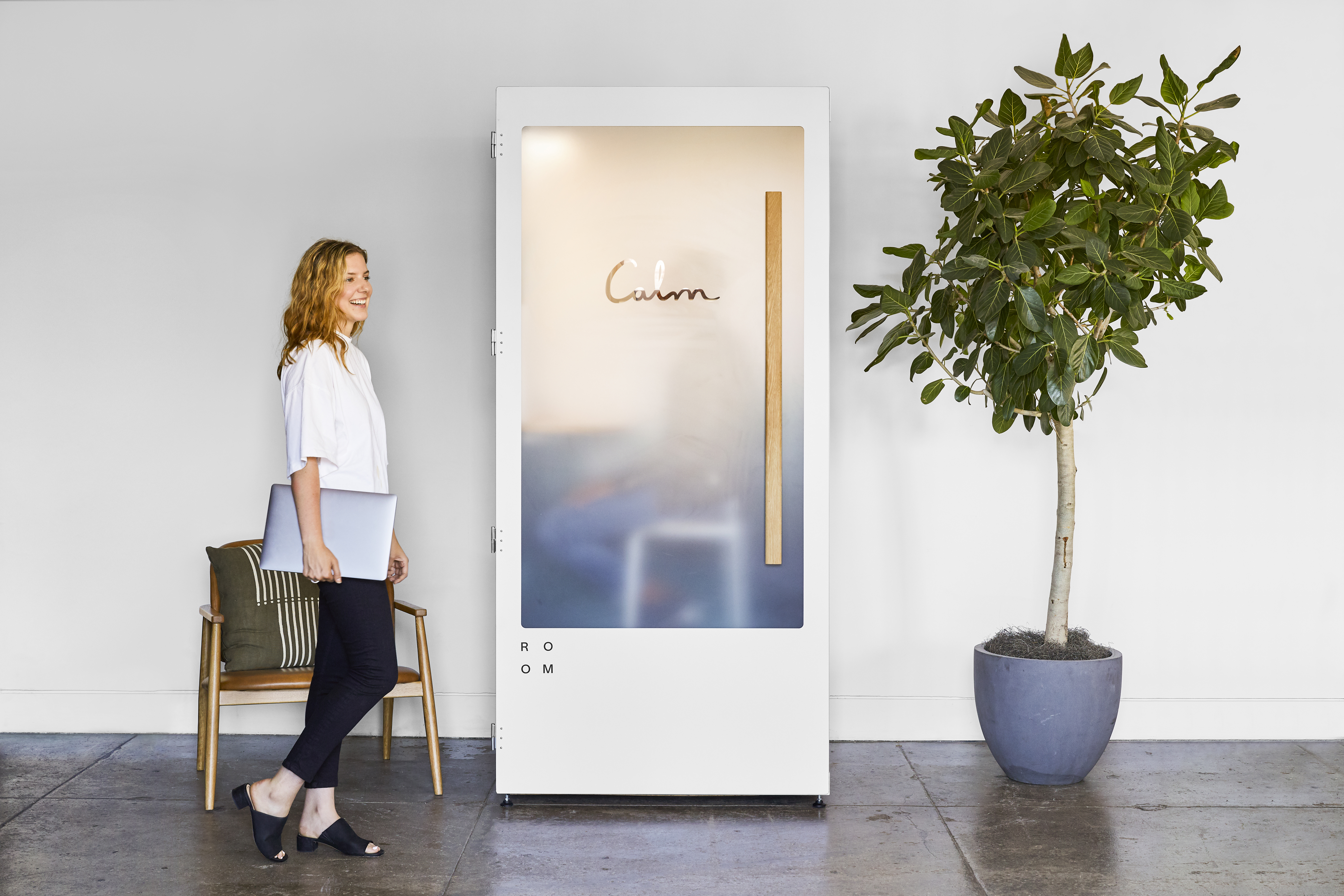 A woman walking toward a Calm meditation booth made by ROOM. The booth is white with a frosted acrylic door.