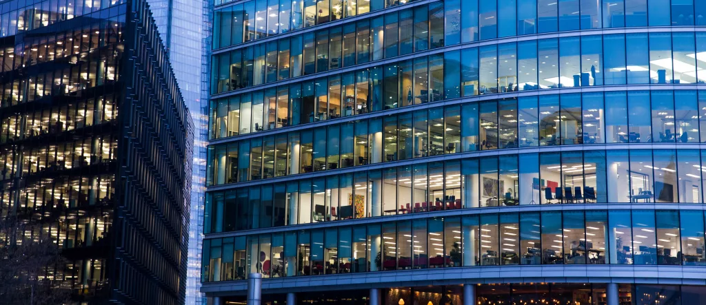 Light shines into the windows revealing seats and desks inside a curved glass high rise. 